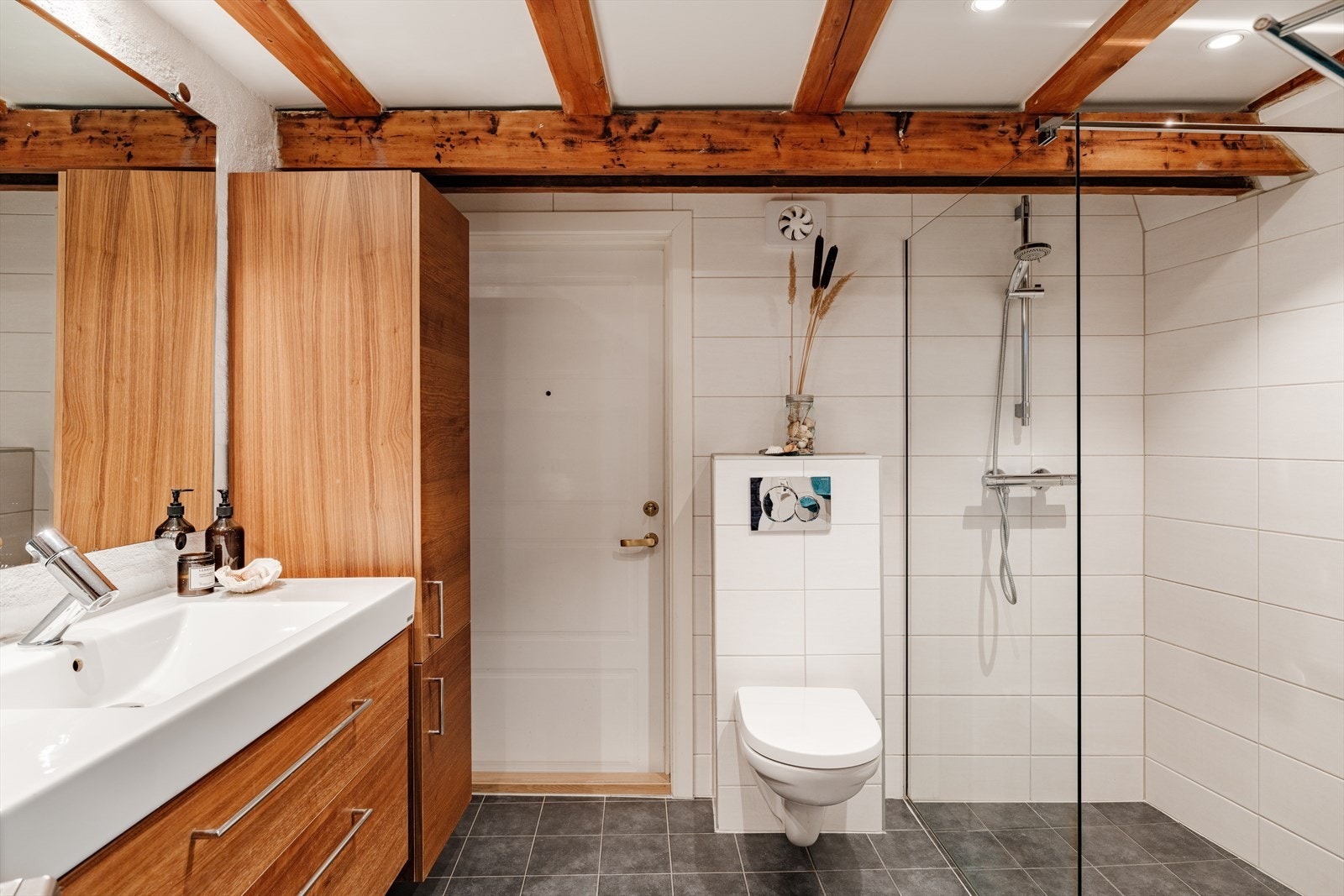 Romslig bad med opplegg for vaskemaskin. Badet har flislagt gulv med gulvvarme, vegger med fliser og malte flater, samt malt tak med synlige trebjelker og downlights. Galleribilde