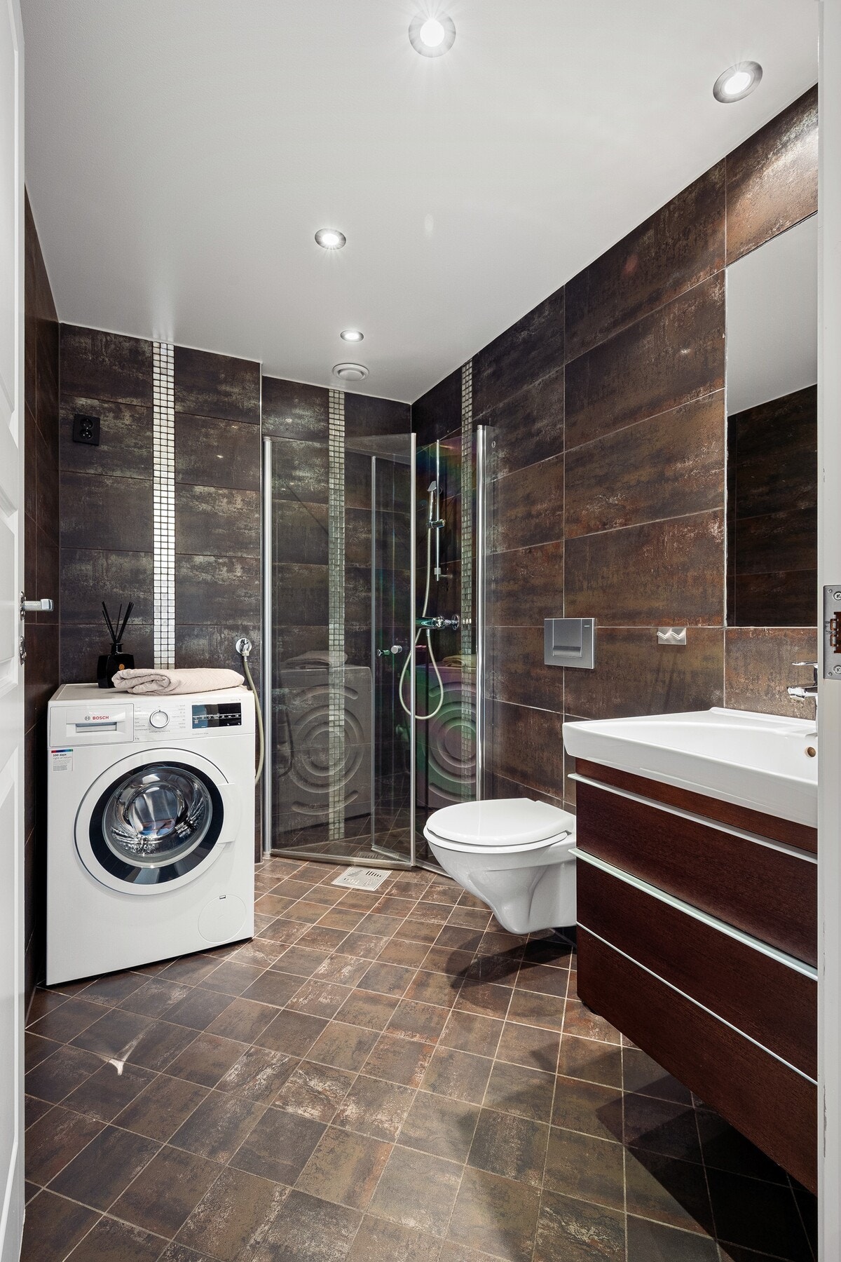 Badet er pent innredet med mørke fliser på overflater. I tillegg finnes det veggmontert toalett, dusjhjørne med glassdører, samt opplegg for vaskemaskin. Galleribilde