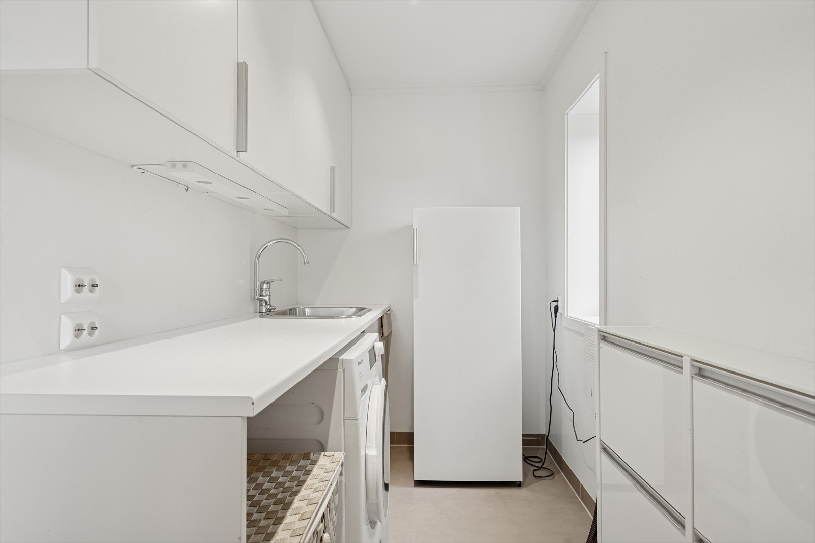 Innenfor badet er det et kjekt vaskerom med flislagt gulv og slette hvitmalte vegger - Rommet har utslagsvask, opplegg for vaskemaskin og romslig innredning med benkeplate og skap Galleribilde