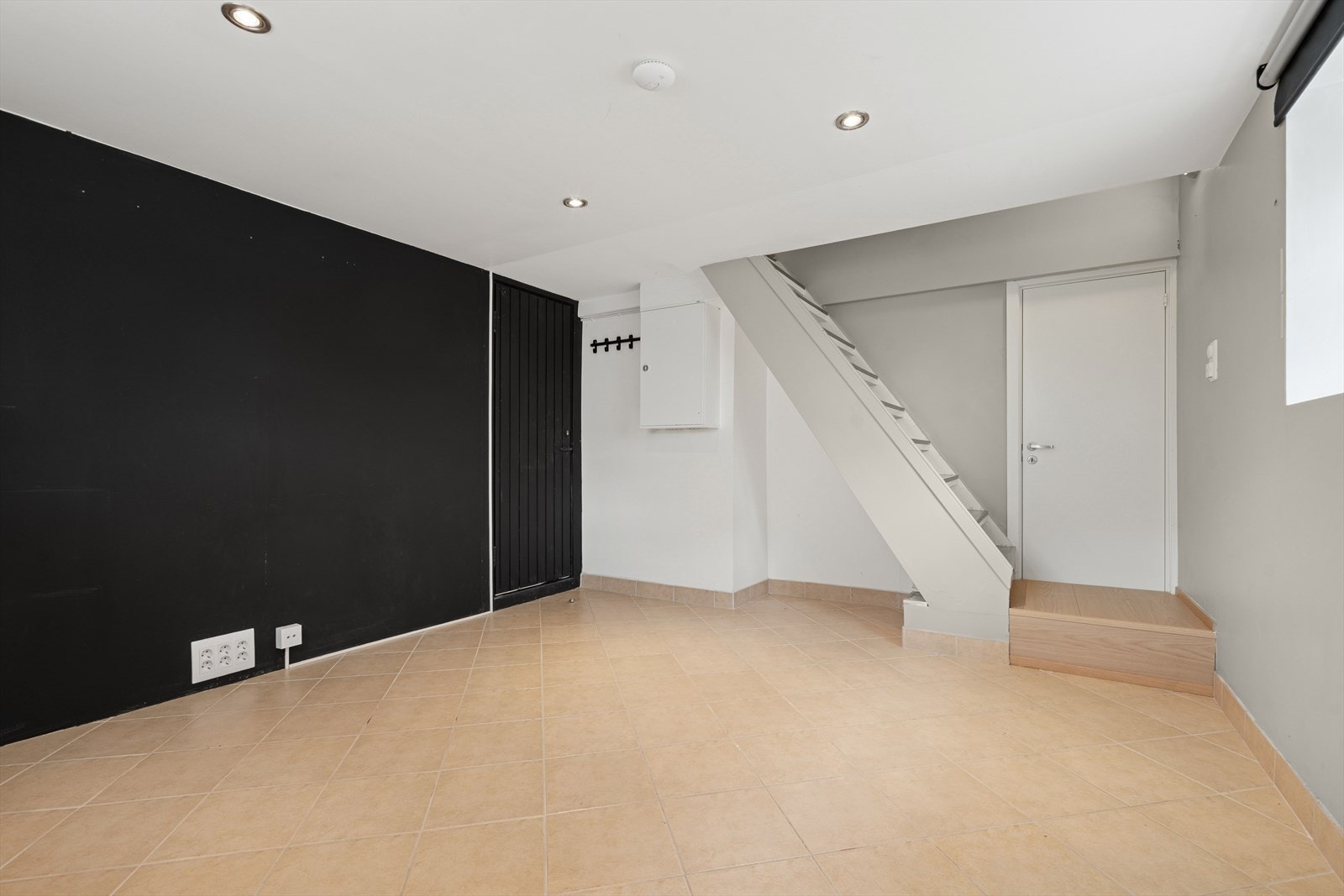 Fra første etasje er det nedgang til underetasjen, hvor du finner du en lys og innbydende kjellerstue - Rommet har flislagt gulv, belysning med downlights, lysmalte vegger og en sort kontrastvegg Galleribilde
