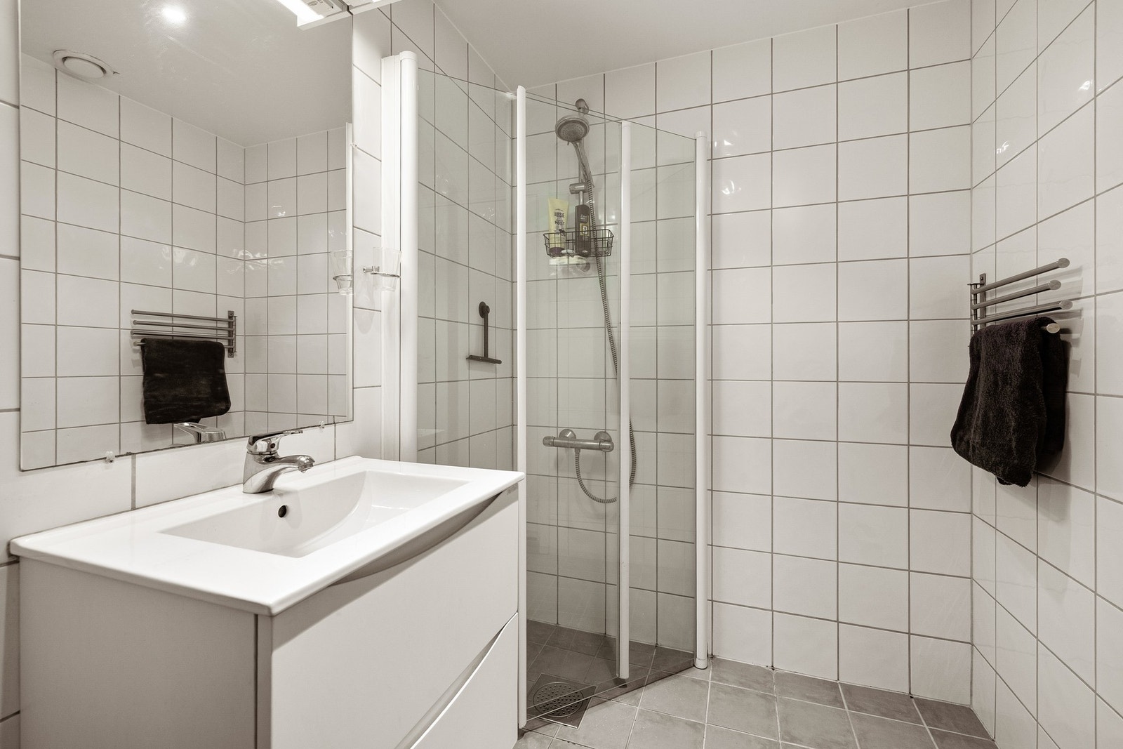 Lys baderomsinnredning med skuffer og heldekkende servant, speil med belysning over. Baderommet er også utstyrt med dusjhjørne med innfellbare glassdører og gulvstående toalett. Galleribilde
