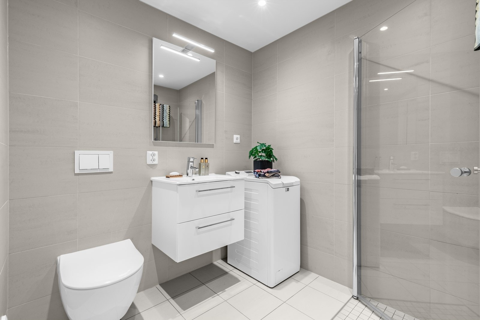 Fra gangen er det inngang til et flislagt og moderne baderom. Badet har dusjhjørne med dører i herdet glass, veggmontert toalett med innebygget sisterne, samt servant med benkeskap under og speil med integrert belysning over. Galleribilde