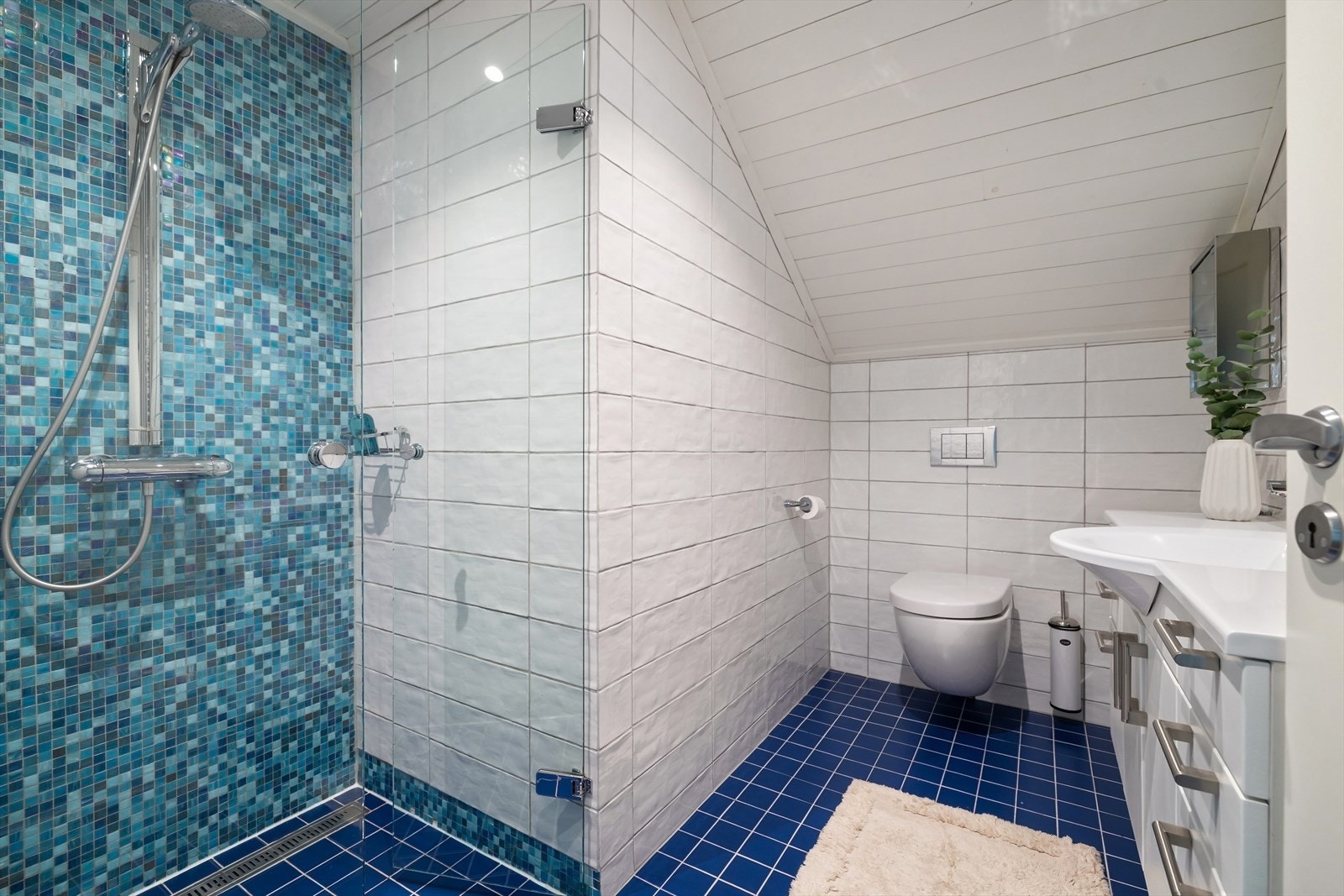 Badet er utstyrt med dusj på gulv, vegghengt toalett og servantinnredning. Galleribilde