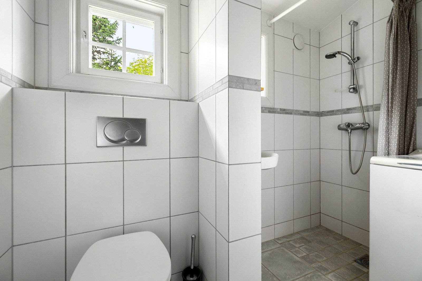 Badet i hybeldelen fremstår som lyst og moderne med flislagte overflater. Innredningen består av servant med underskap, speil, dusj med forheng og veggmontert toalett med innebygget sisterne. Det er også opplegg for vaskemaskin. Galleribilde