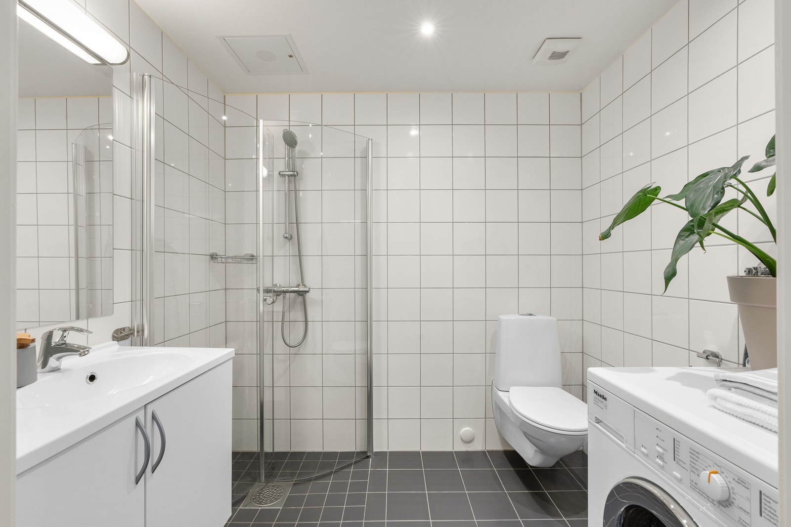 Leiligheten har et lyst og moderne baderom fra byggeåret 2015. Baderommet har mørke fliser på gulv med gulvvarme, hvite fliser på veggene, downlights i himlingen og avtrekksventil. Galleribilde