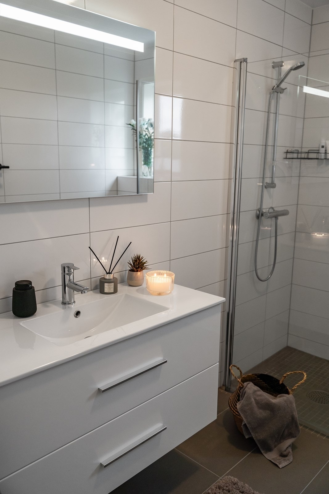 Badet er innredet med hvit baderomsinnredning, speil med belysning, vegghengt toalett samt dusjhjørne med mosaikkfliser og to glassdører. Galleribilde