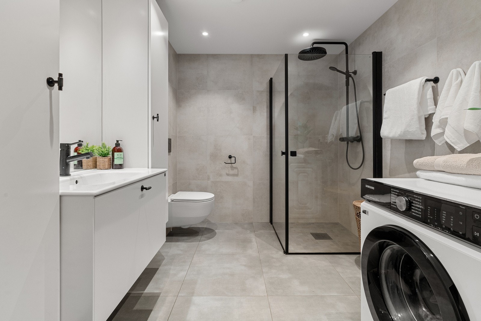 Lekkert baderom med fliser i gulv og vegger, badet er utstyrt med vegghengt servant med oppbevaring, dusjhjørne, samt opplegg til vaskemaskin som gir enkelhet i en travel hverdag. Galleribilde