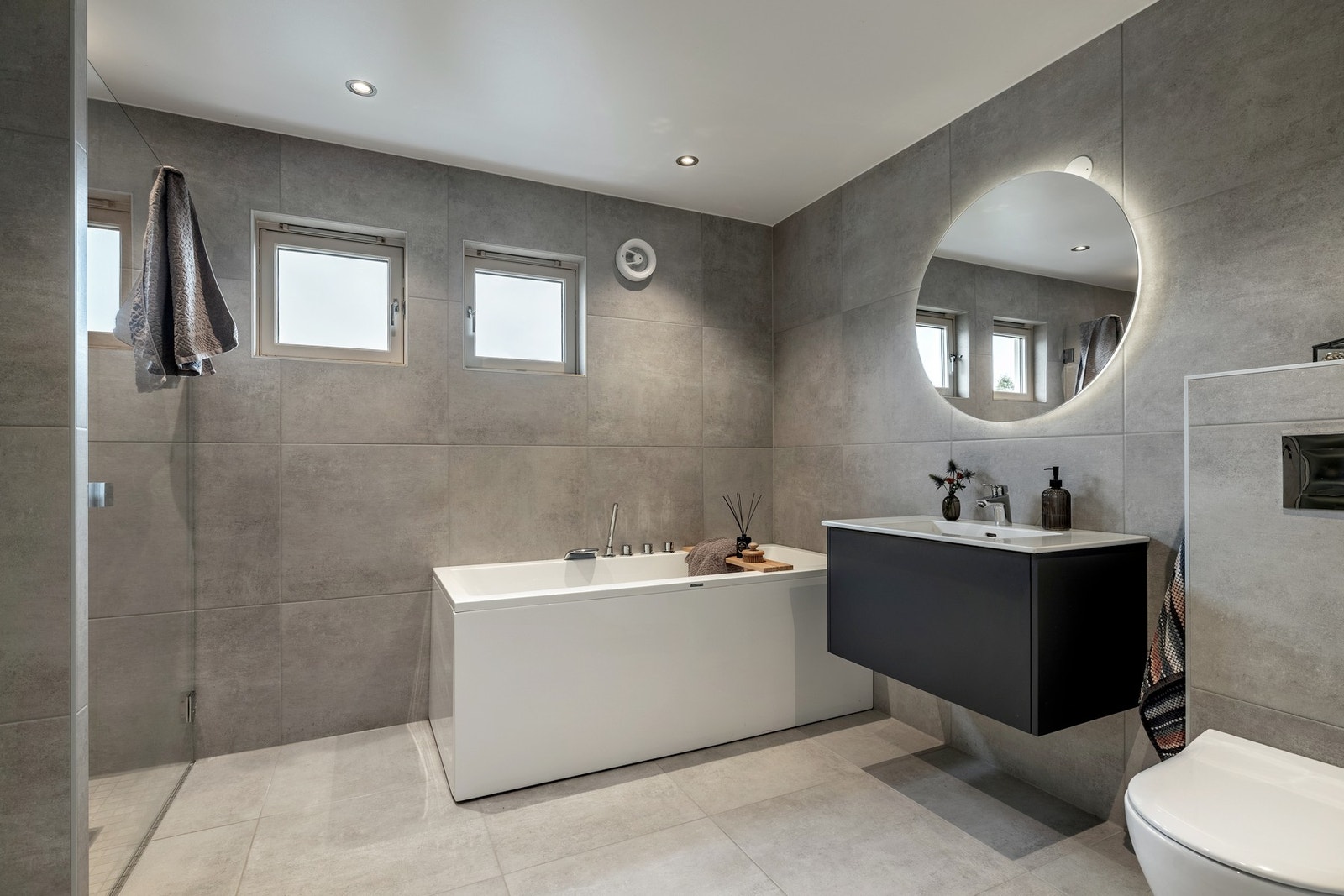 Badet er fra 2024, og fremstår stilfullt og moderne. Badet er utstyrt med store vegg- og gulvfliser i grå nyanser, elektriske varmekabler og innfelte downlights. Galleribilde