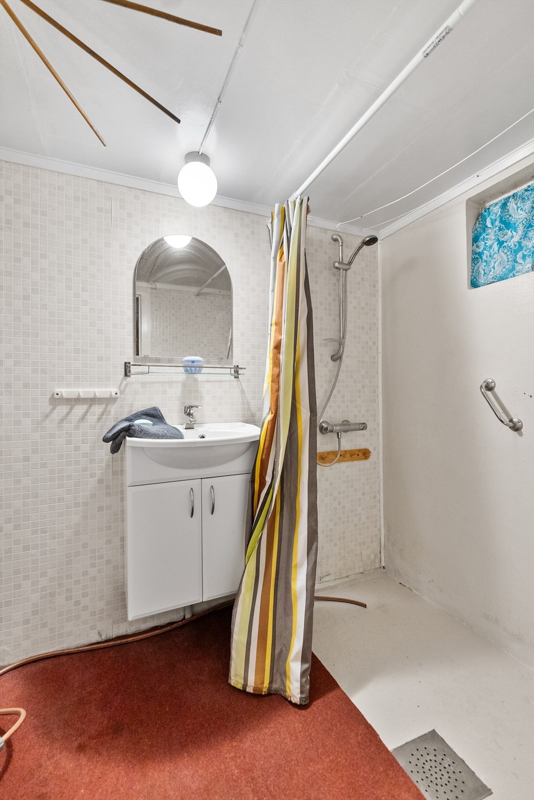 Badet i kjelleren er av eldre dato. Gulvet er av malt betong og teppe, veggene er i malt mur og belegg/tapet, og taket har malte plater. Rommet er innredet med dusjnisje med forheng, servant i innredning og speil. Galleribilde