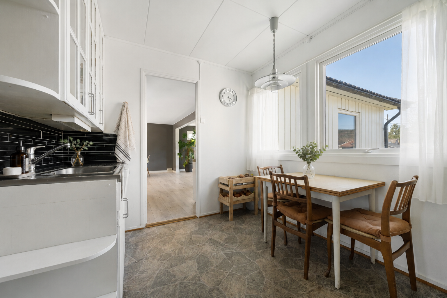 Fra stuen er det tilkomst til et lyst og trivelig kjøkken med store vindusflater, og plass til spisebord. Deler av bildet er møblert med KI. Galleribilde