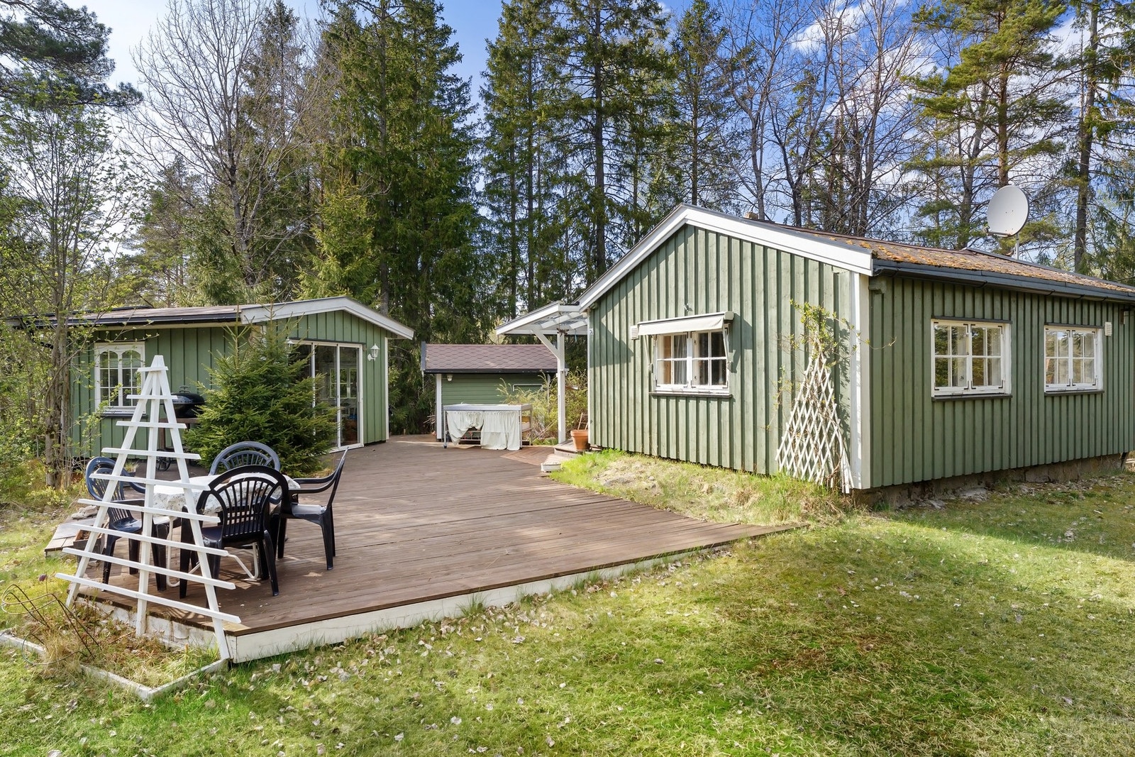 Grønnmalt fritidsbolig med trebygninger, hagestue, redskapsbod og stor terrasse, omgitt av skog. Galleribilde