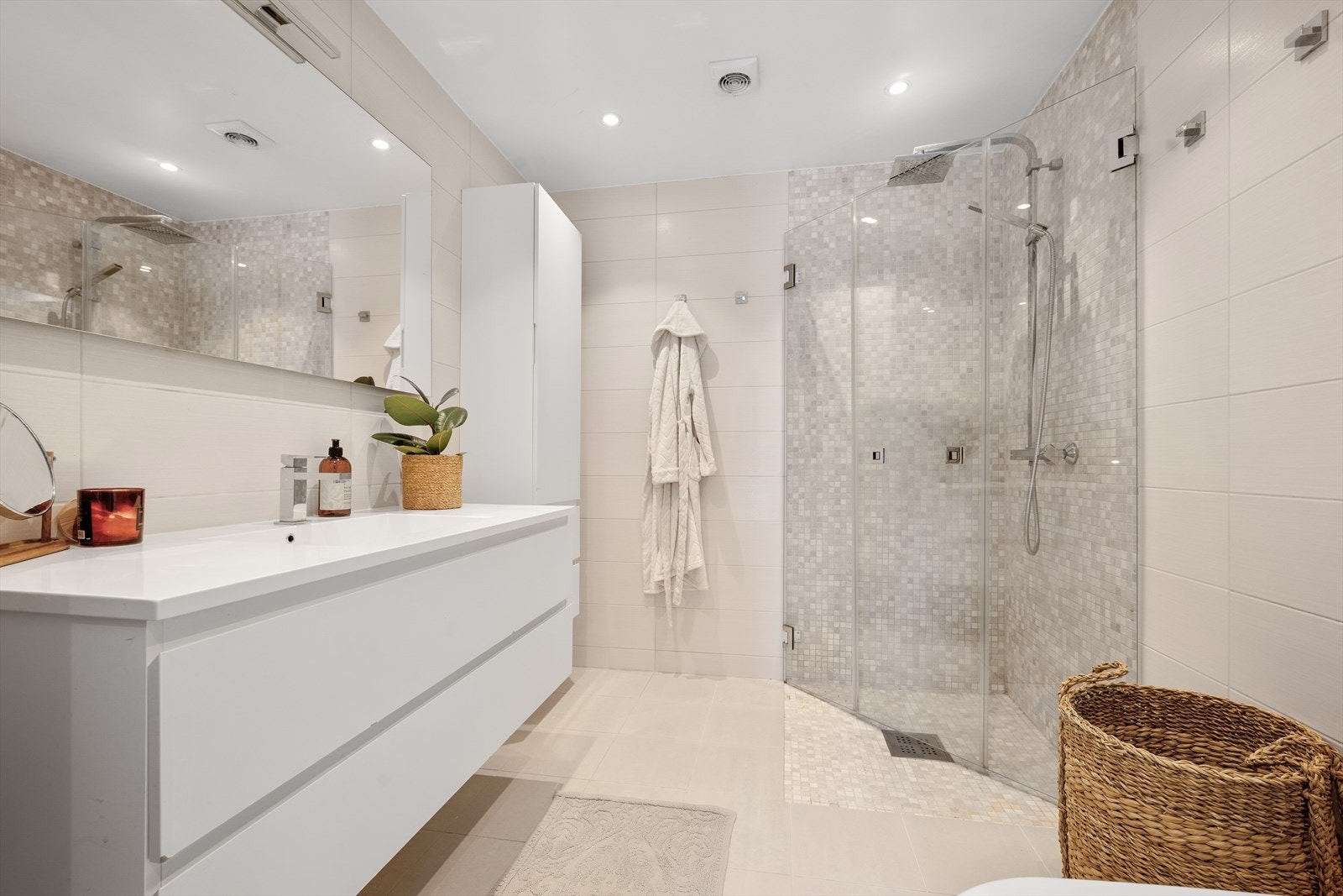 Badet har en gjennomgående palett av lyse, varme beige- og kremtoner som gir et rolig og innbydende uttrykk. Veggene er kledd med store, matte fliser i en myk sandfarge, mens dusjsonen fremheves av små mosaikkfliser i varierte nyanser av lys beige og grå. Galleribilde