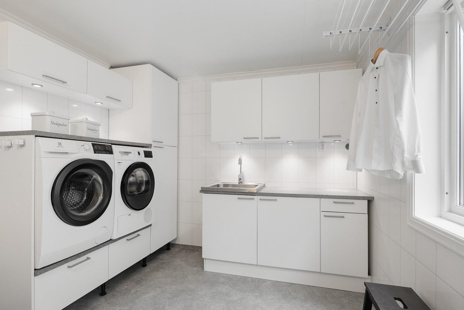 Vaskerom med vinylbelegg på gulv og hvite baderomsplater på vegg. Kjekk og praktisk vaskeromsinnredning med hevet plass til vaskemaskin og tørketrommel. Grå benkeplate med vask i stål. Galleribilde