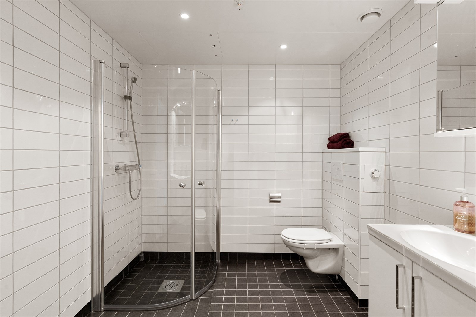 Ett av leilighetens 2 bad. Pent, helfliset bad/vaskerom. Hvit seksjon med vask/benk og speilskap på vegg. Dushjørne og vegghengt toalett. Opplegg for vaskemaskin. Galleribilde