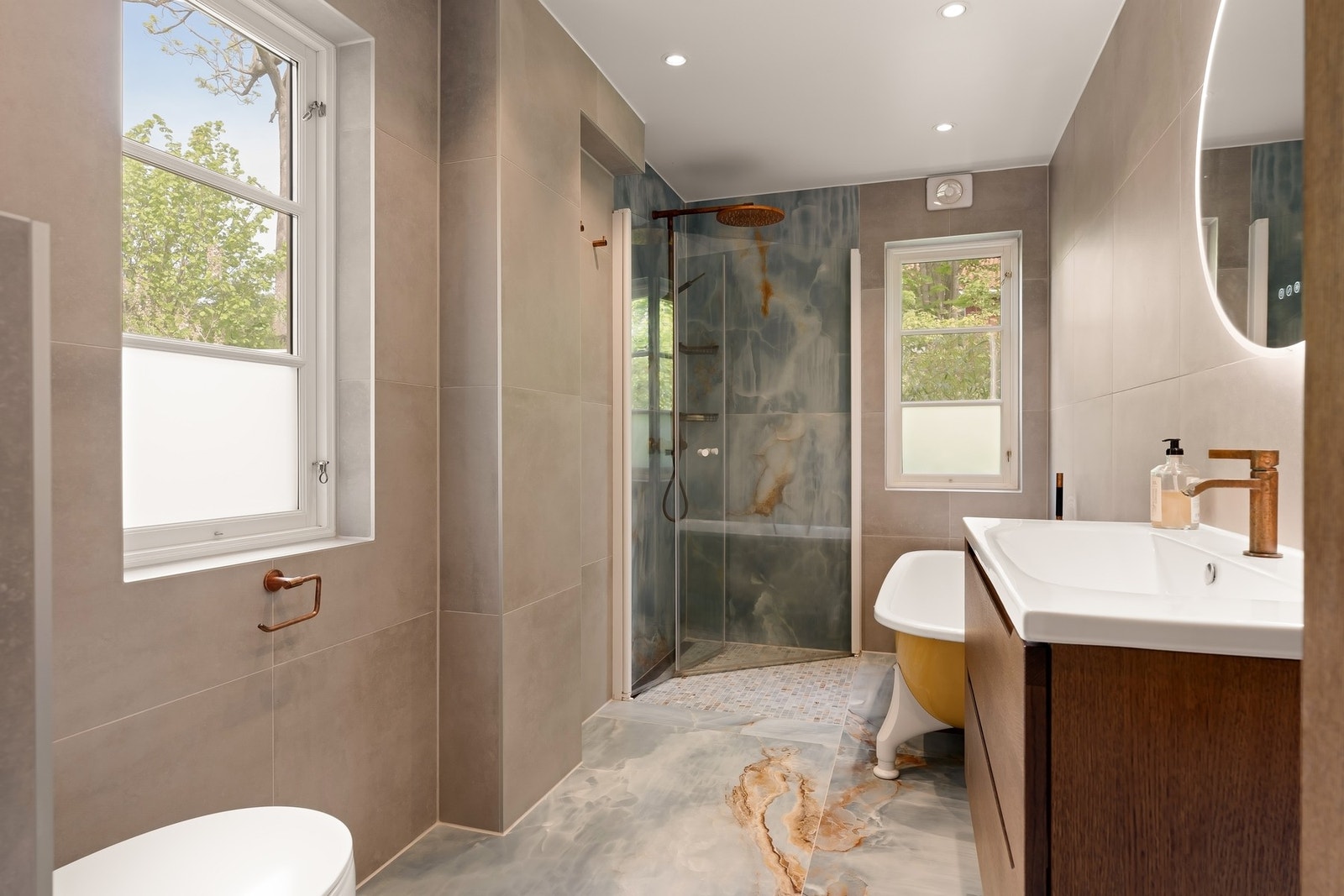 Badet er utstyrt med veggmontert wc, baderomsinnredning, badekar, dusjhjørne og speil med integrert belysning Galleribilde