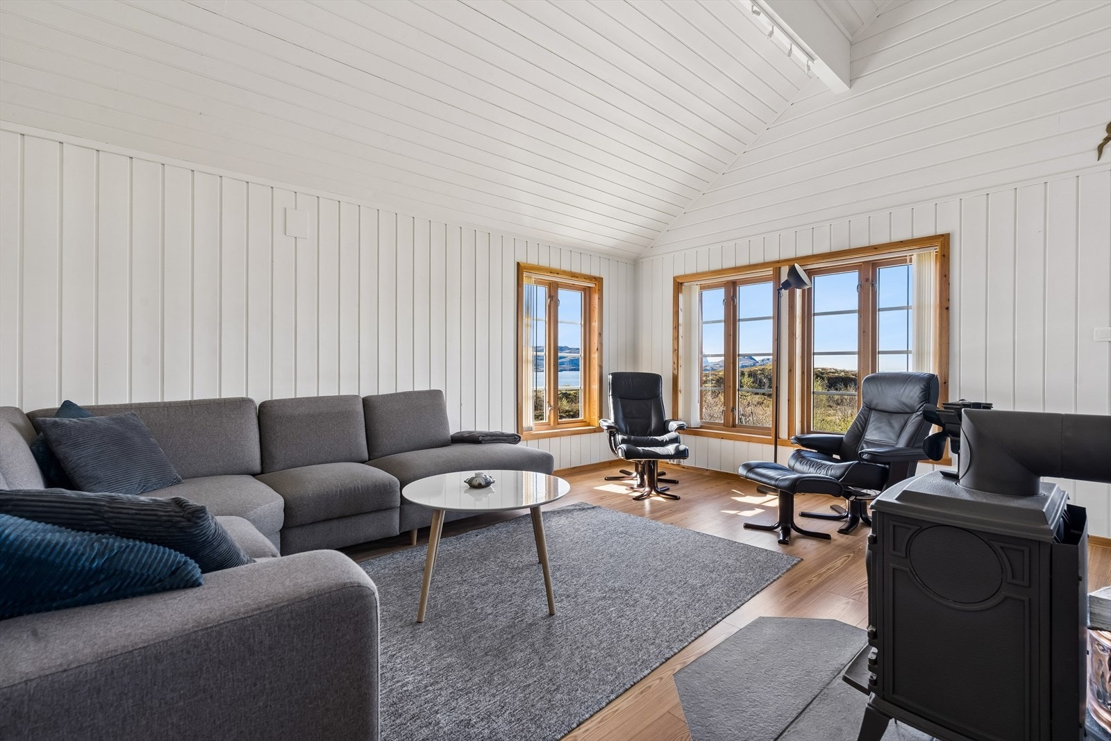 Lyst og luftig allrom med lysmalte panelvegger og vedovn. Direkte utgang til terrassen visker ut skillet mellom ute og inne. Galleribilde