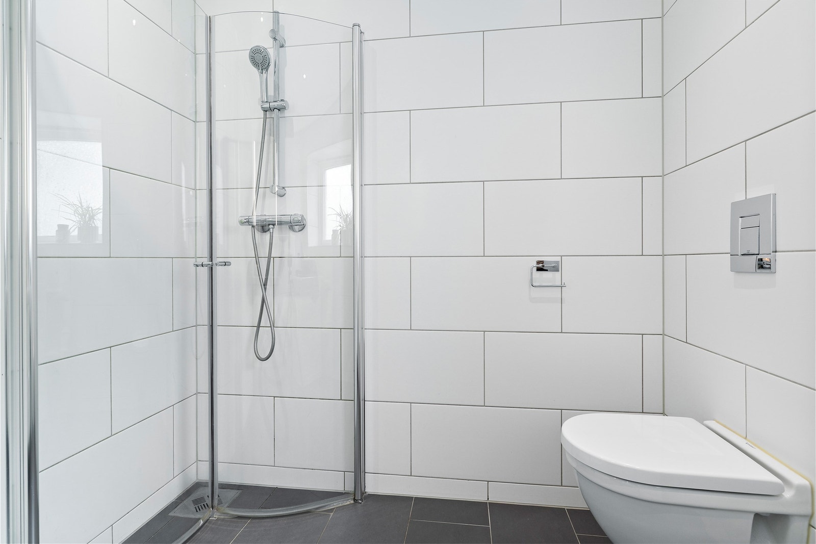 Dusjhjørnet har plassbesparende, foldbare dører og stilren armatur. Videre er det veggmontert toalett, opplegg for både vaskemaskin og tørketrommel, samt mekanisk avtrekk fra badet Galleribilde