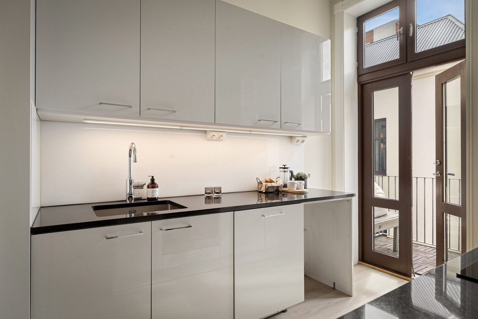 Blum kjøkkeninnredning med glatte fronter og benkeplate av stein med underlimt vaskekum. Frostet glassplate og belysning over benkeplaten Galleribilde