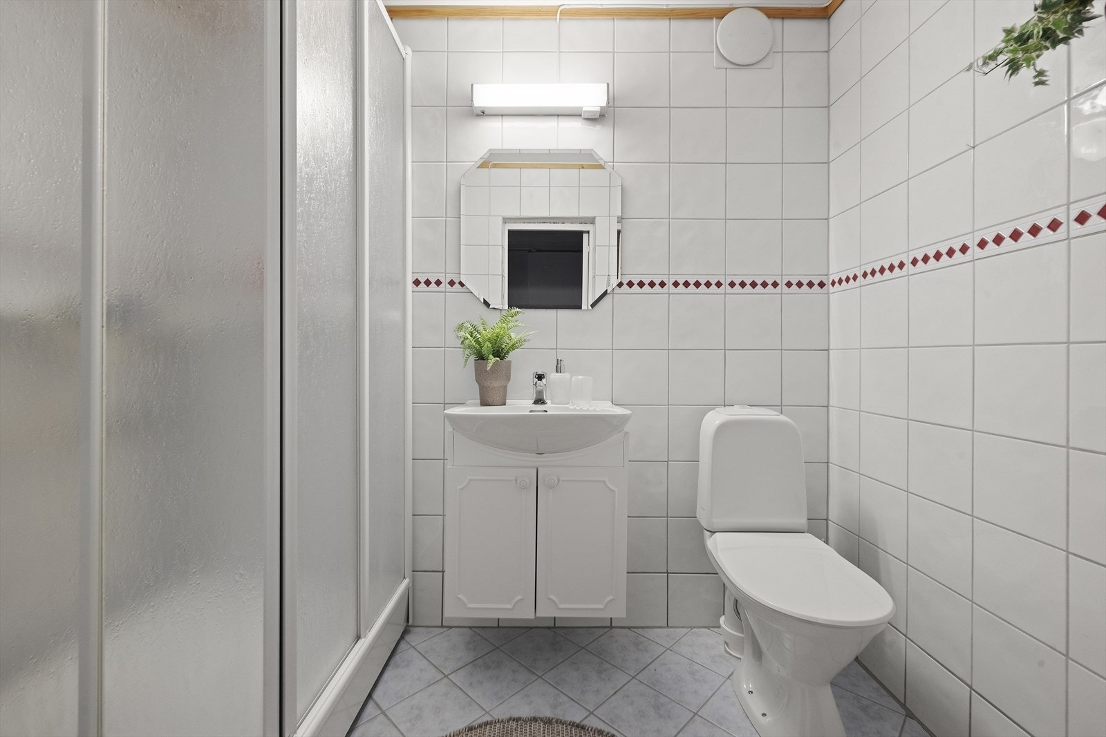 Badet i underetasjen er tidløst innredet med lyse fliser på gulv og vegger, og er ellers utstyrt med dusjkabinett, toalett og baderomsinnredning med servant. Kabinettet har produksjonsår i 1999, noe som kan indikere at badet er fra rundt denne tiden. Galleribilde