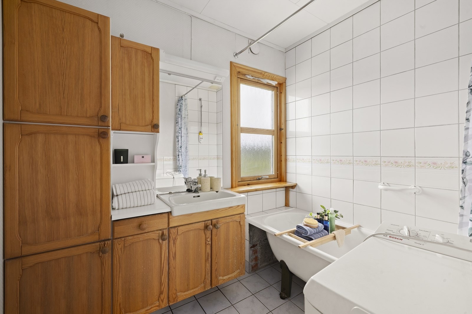 Badet er innredet med lyse fliser på gulvet og en kombinasjon av lyse fliser og tapetserte plater på vegg. Badet er ellers utstyrt med badekar med dusj og baderomsinnredning med god lagringsplass og nedfelt servant. Galleribilde