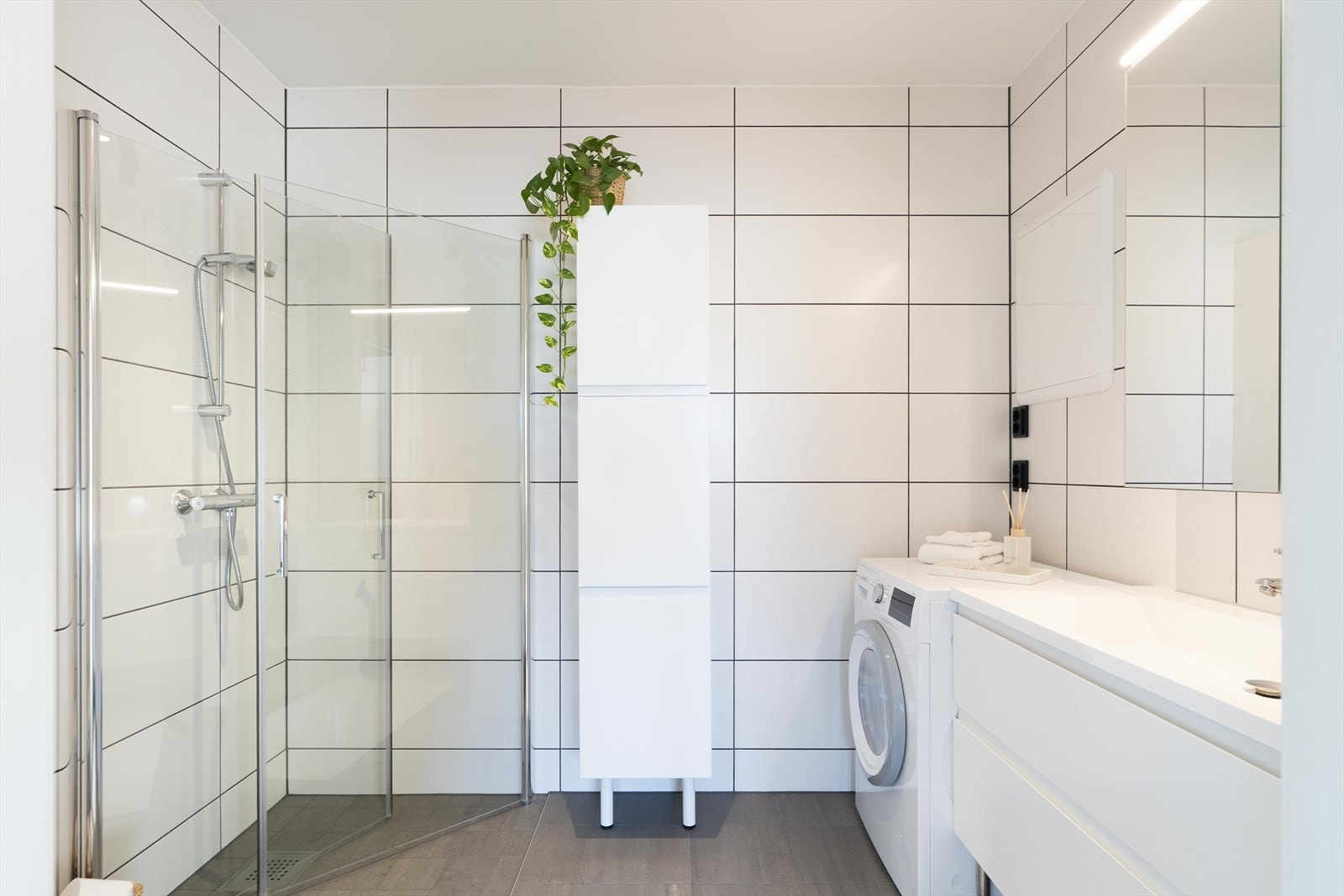 Badet fremstår lyst og moderne, med store hvite fliser på vegg og mørkere flislagt gulv. Rommet er innredet med dusjsone med glassvegger, servantinnredning med skuffer og speil med belysning. Galleribilde