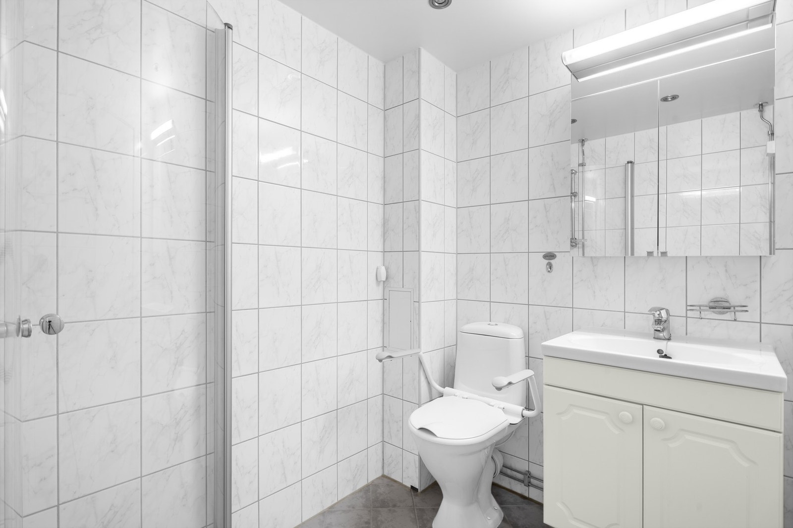 Videre er badet utstyrt med servantskap med profilerte fronter og heldekkende servant, gulvmontert klosett, dusjhjøne med svingbare dører i glass og blandebatteri. Galleribilde