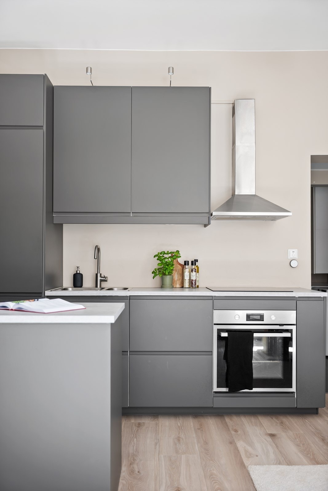 Kjøkkenet har en moderne innredning med grå, glatte fronter og hvit laminatbenkeplate med nedfelt kum i rustfritt stål. Galleribilde