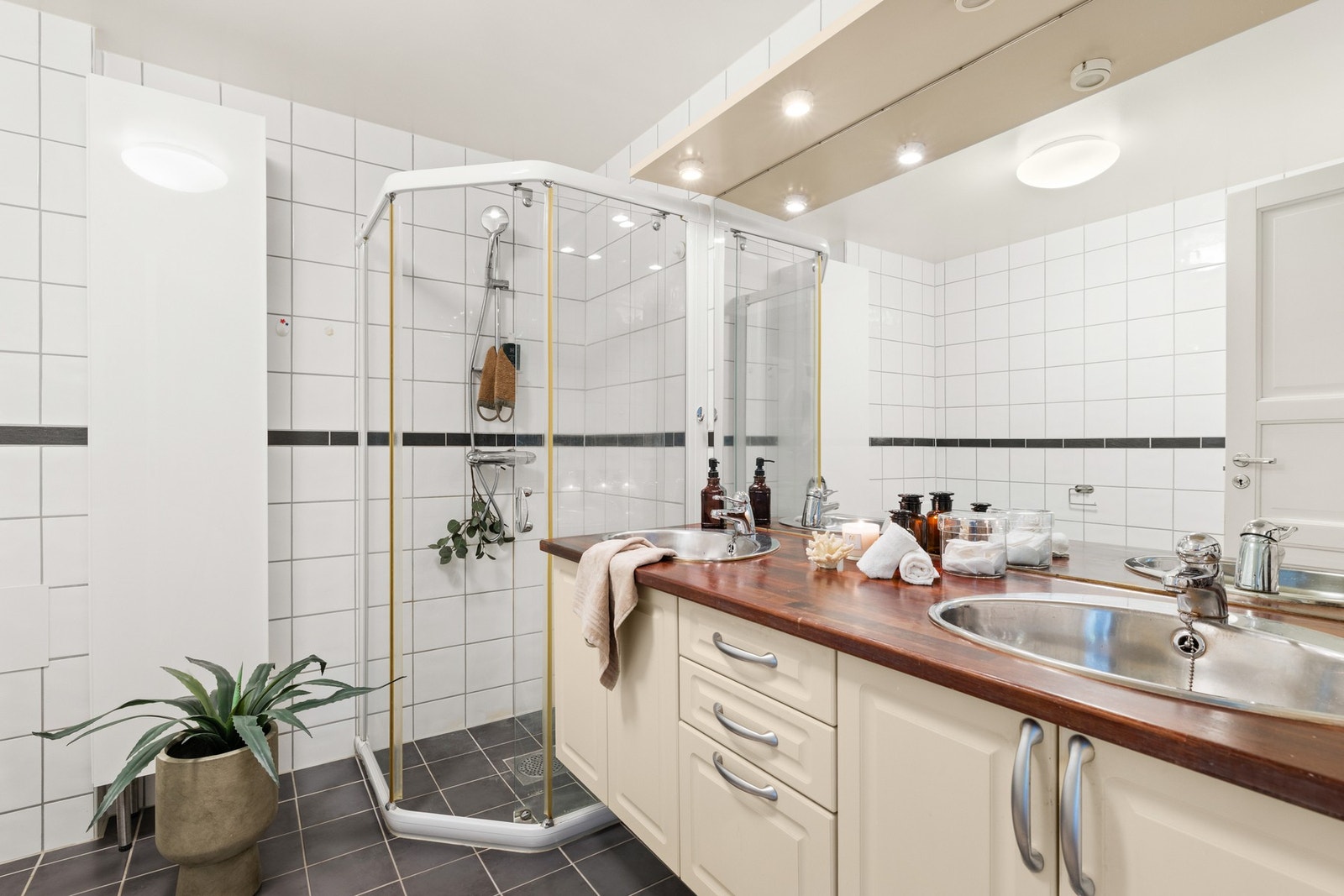Badet er flislagt på gulv og vegger, og har malte takflater med belysning. Rommet er innredet med veggmonterte dusjvegger, dusj direkte på gulv med termostatbatteri, klosett og dobbel servant med skapinnredning. Galleribilde