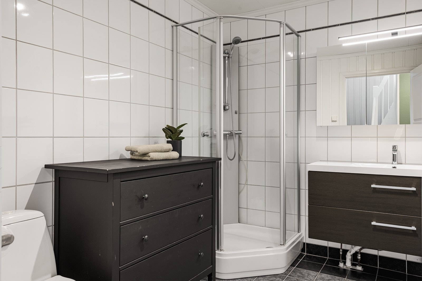 Badet er utstyrt med dusjkabinett, gulvmontert toalett og baderomsinnredning med skuffer og heldekkende servant med speilskap i overkant. Galleribilde