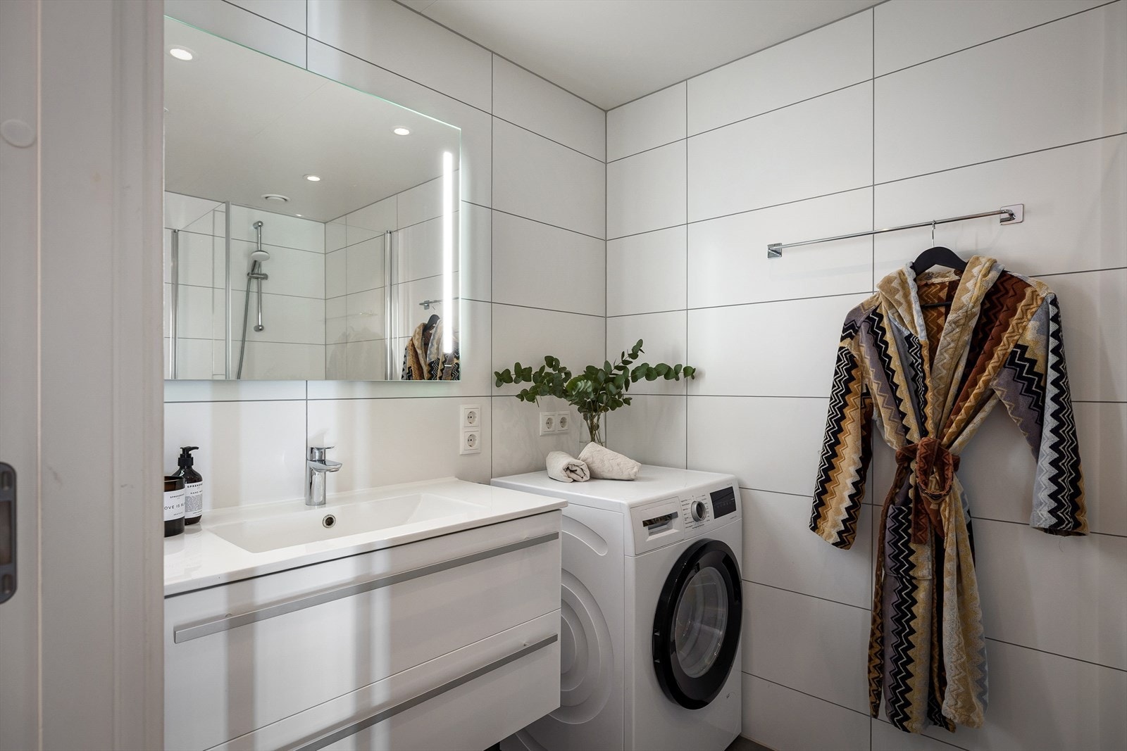 Badeværelset er romslig med hvite vegger og grått gulv, med dusjsone og opplegg for vaskemaskin. Ovenpå vaskemaskinen får du enkelt satt en tørketrommel. Galleribilde