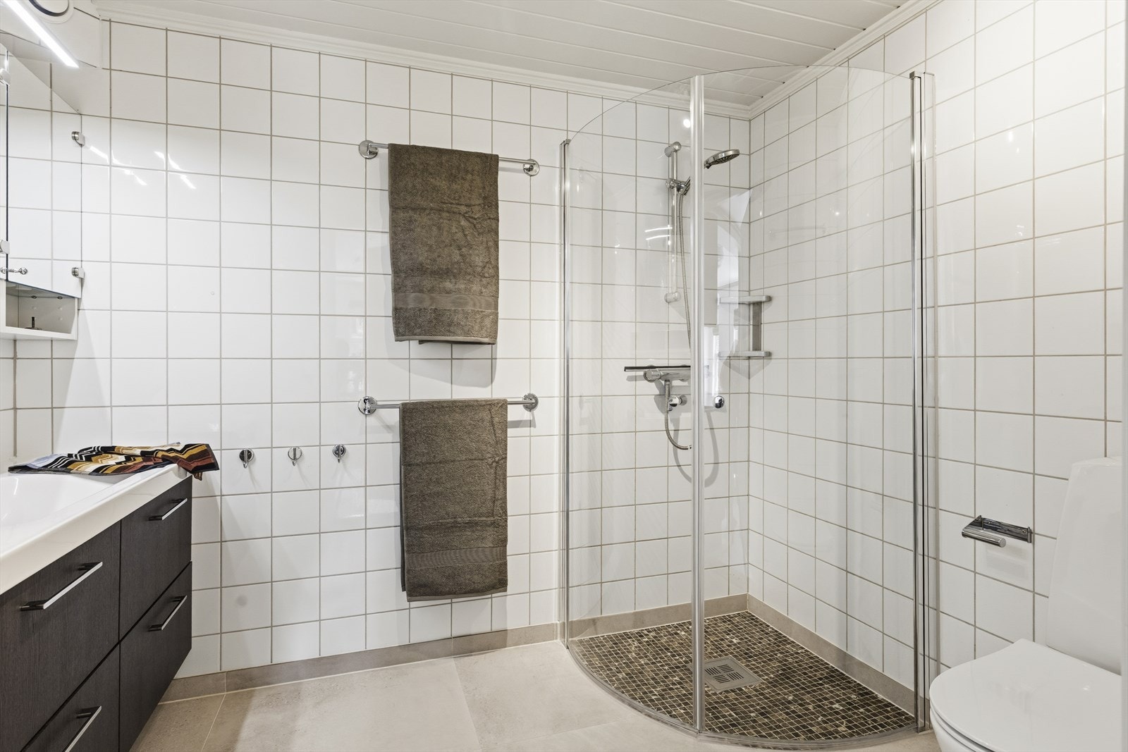 Badet er innredet med vegghengt toalett, dusjhjørne med glassdører og stor baderomsinnredning med speilskap. Opplegg for vaskemaskin Galleribilde