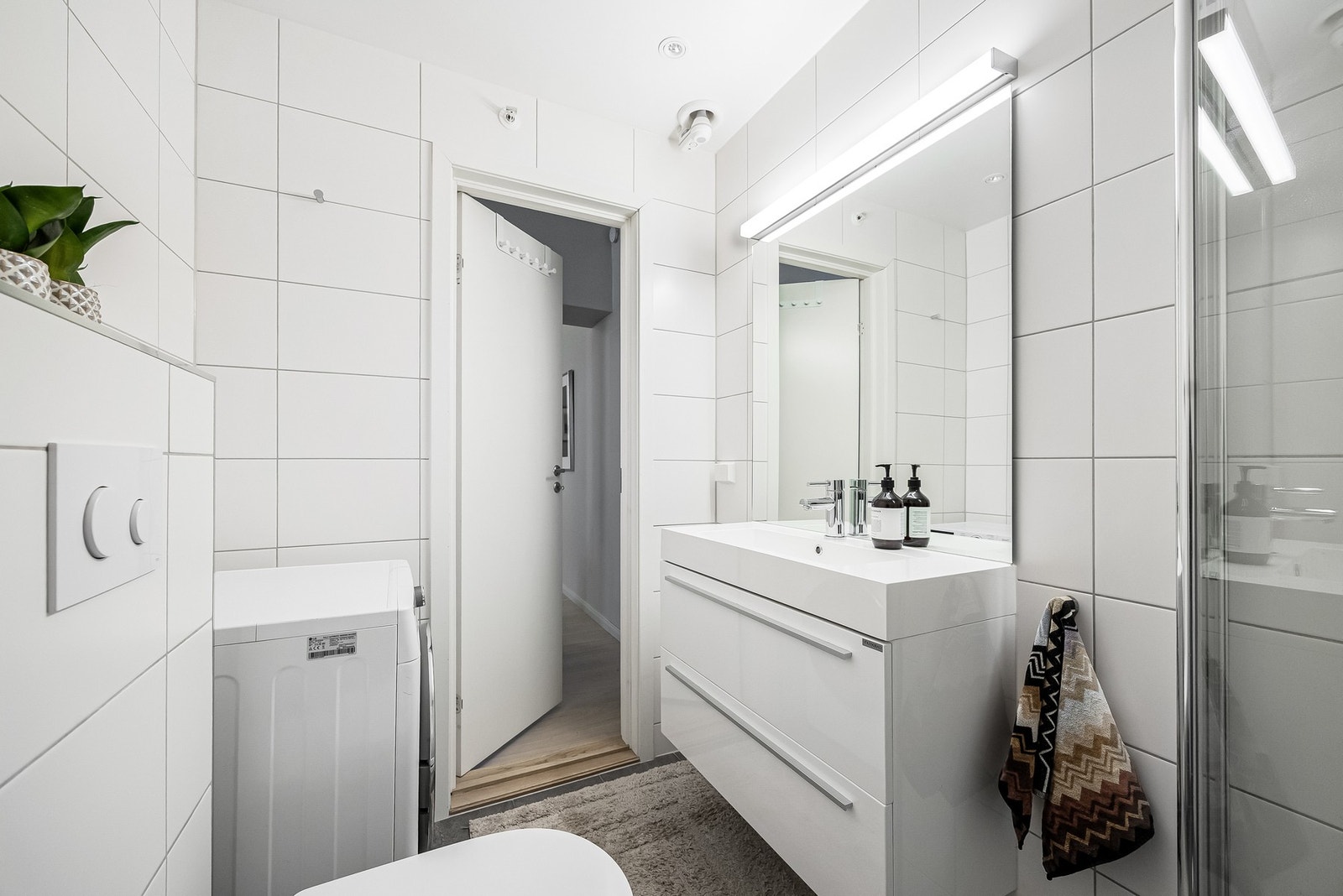 Badet har dusj med innfellbare dusjdører i herdet glass, servant i helstøpt plate, vegghengt toalett med innebygget sisterne samt opplegg for vaskemaskin. Galleribilde