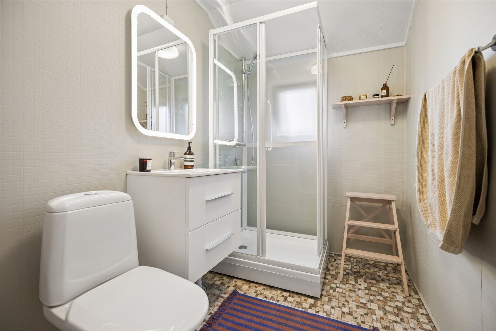 Bad med fliser på gulv med varme. Badet er i hovedsak fra byggeår, med noen oppgraderte overflater. Nyere dusjkabinett, servantinnredning, speil og toalett. Galleribilde