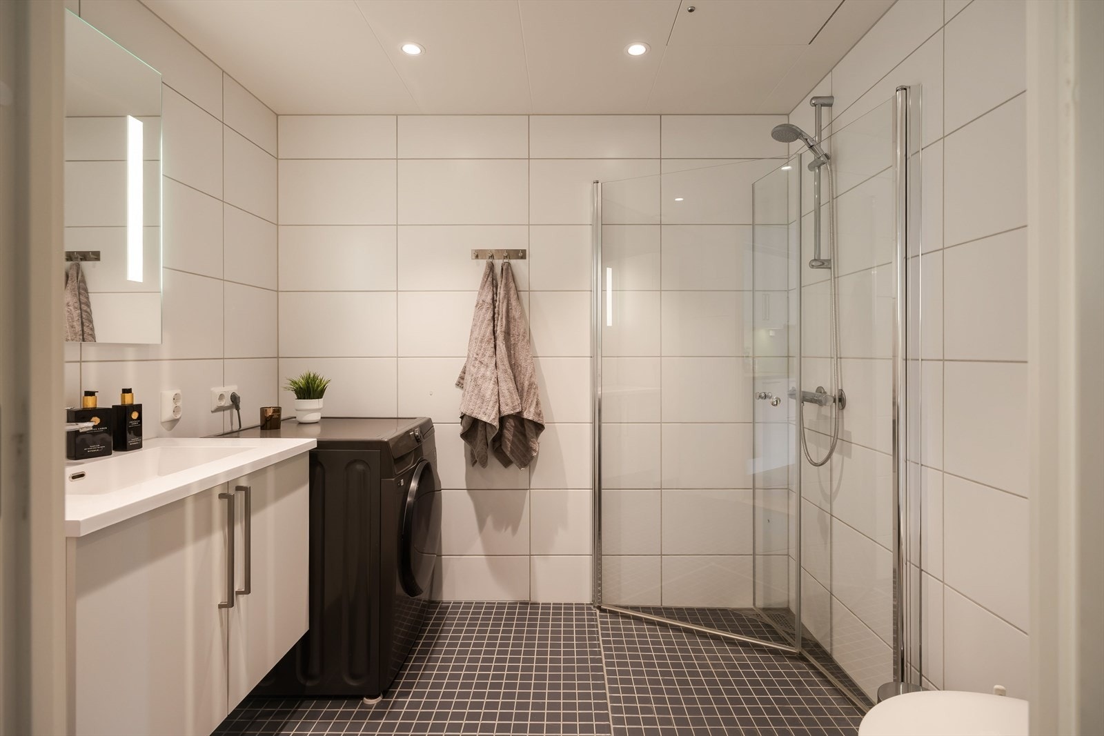 Badet består av gulv- og veggfliser, innfellbare dusjvegger av glass, vegghengt toalett, servantskap, lysspeil og opplegg for vaskemaskin. Galleribilde