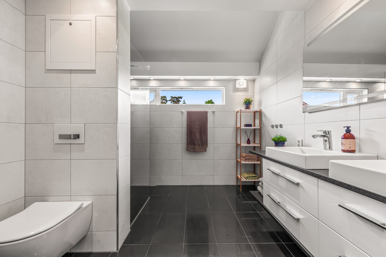 Badet er innredet med store vegg- og gulvfliser i ulike grånyanser. Varme er lagt i gulvet, og badet har downlights, elektrisk sensorvifte og vegghengt toalett. Galleribilde
