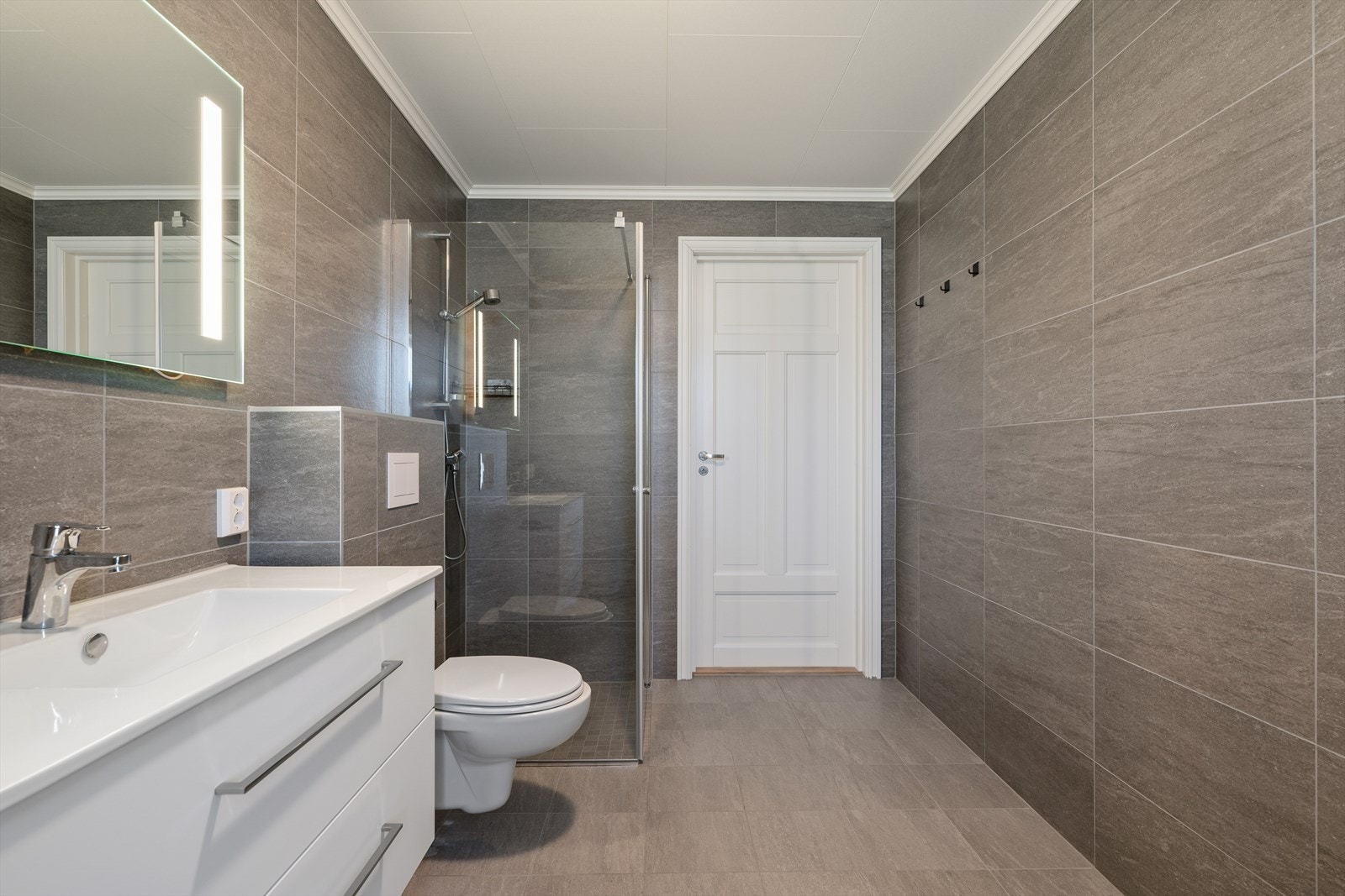 Badet er innredet med hvit baderomsinnredning med nedfelt servant, slette lyse fronter og speil med integrert belysning. Galleribilde