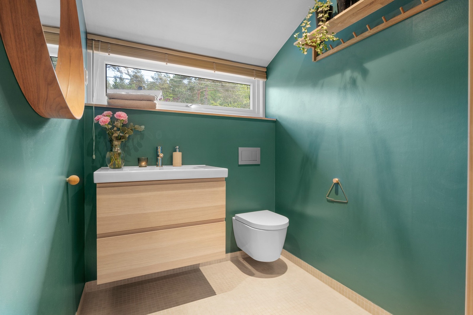 Fra motsatt side kommer det daglys inn fra et vindu plassert høyt på veggen. Innredningen består av en heldekkende servant på trehvit skuffeseksjon, speil og veggmontert toalett. Galleribilde