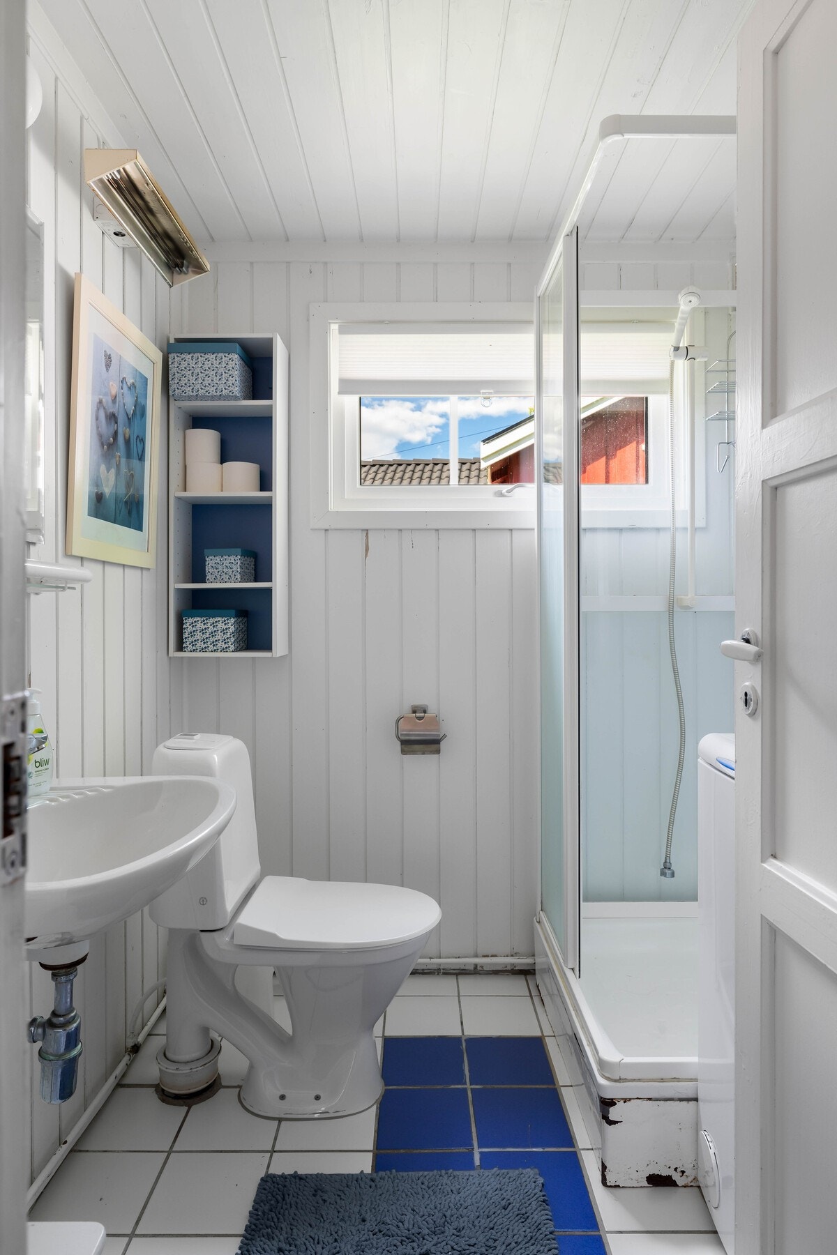 Badet har hvite og blå gulvfliser, hvitmalte panelvegger og hvitmalt panelhimling. Rommet er innredet med et dusjkabinett, toalett, servant, speil og hyller. Galleribilde