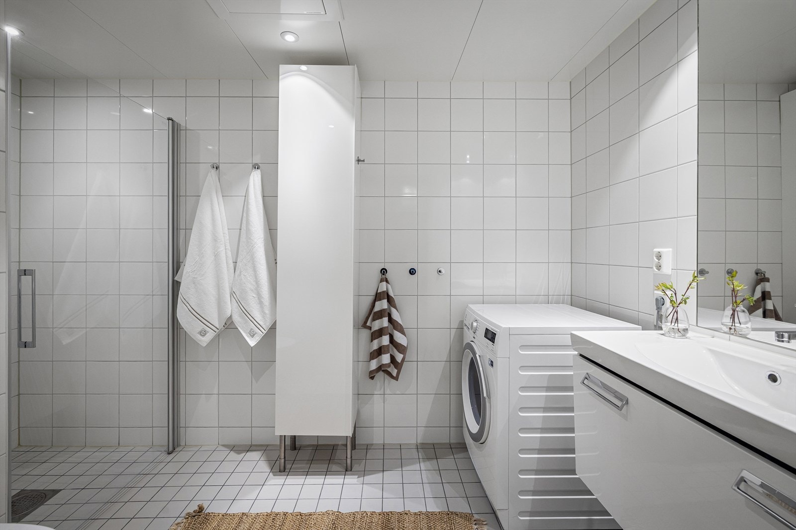 Badeværelset er flislagt hvitt, noe som føles rent og svalt. Her er en stilren dusjsone og opplegg for vaskemaskin. Galleribilde