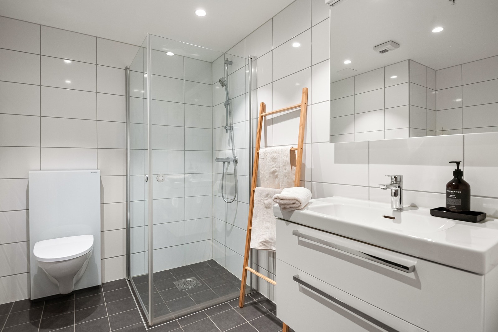 Leiligheten har et lyst og moderne baderom fra byggeåret 2020. Baderommet har mørke fliser på gulv, elektrisk gulvvarme, hvite fliser på veggene og downlights i himlingen. Galleribilde