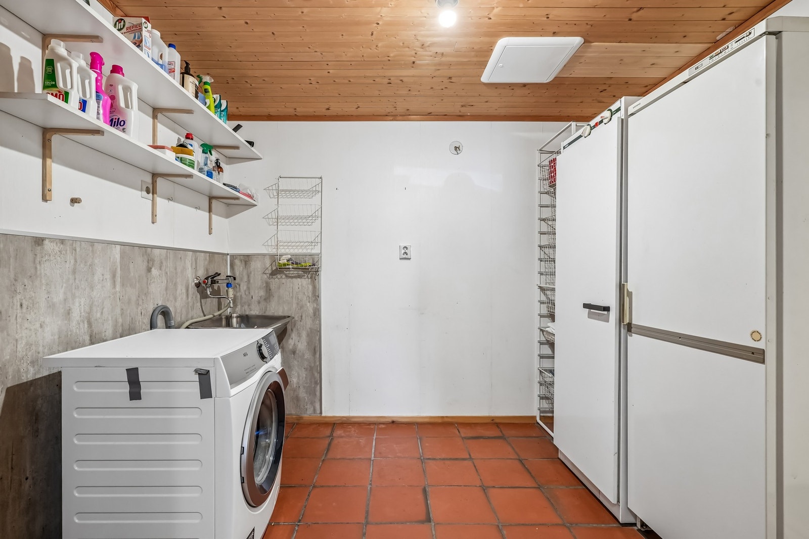 Praktisk vaskerom med klinkerfliser på gulvet og tapet på vegger, det er våtromsplater på vegg bak vaskemaskin og stålskyllekum. Galleribilde