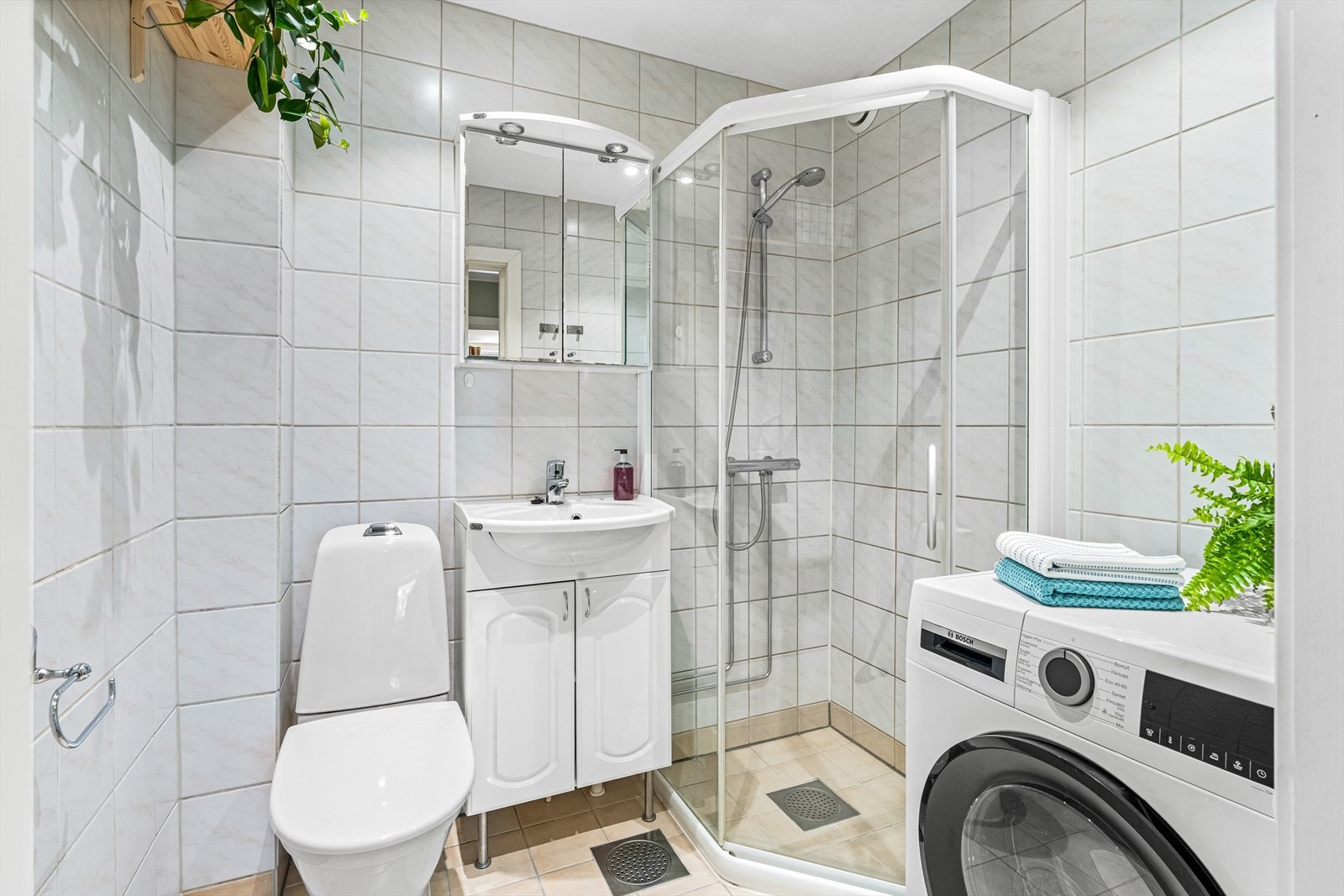 Badet har lyse fliser på gulv og vegger, samt gulvvarme. Innredningen består av et underskap/overskap, servant, speil, lys, stikk og et dusjhjørne. Galleribilde