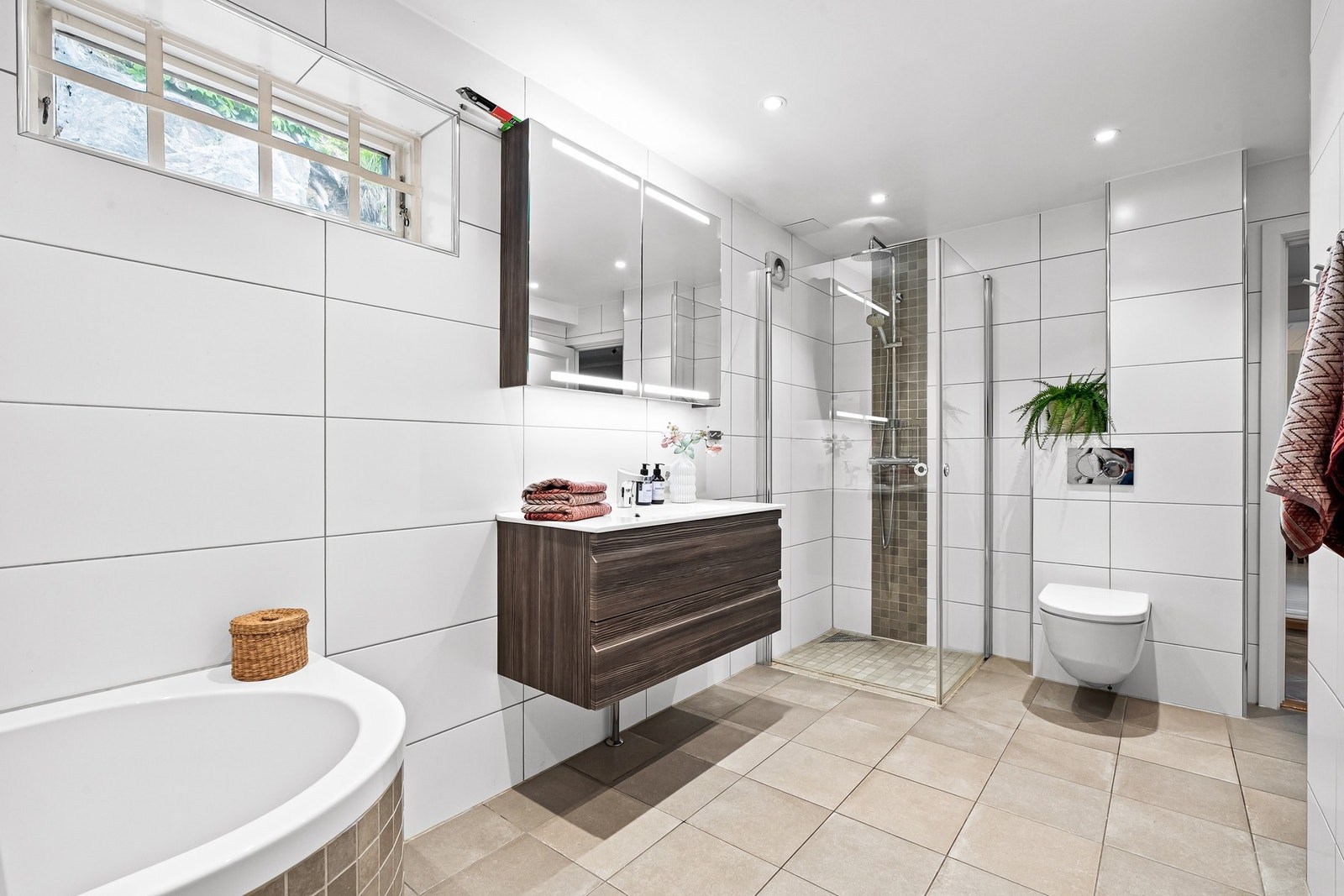 Badet er innredet med vegghengt toalett, sensor vifte, et innfliset badekar og et dusjhjørne med regnfallsdusj. Galleribilde