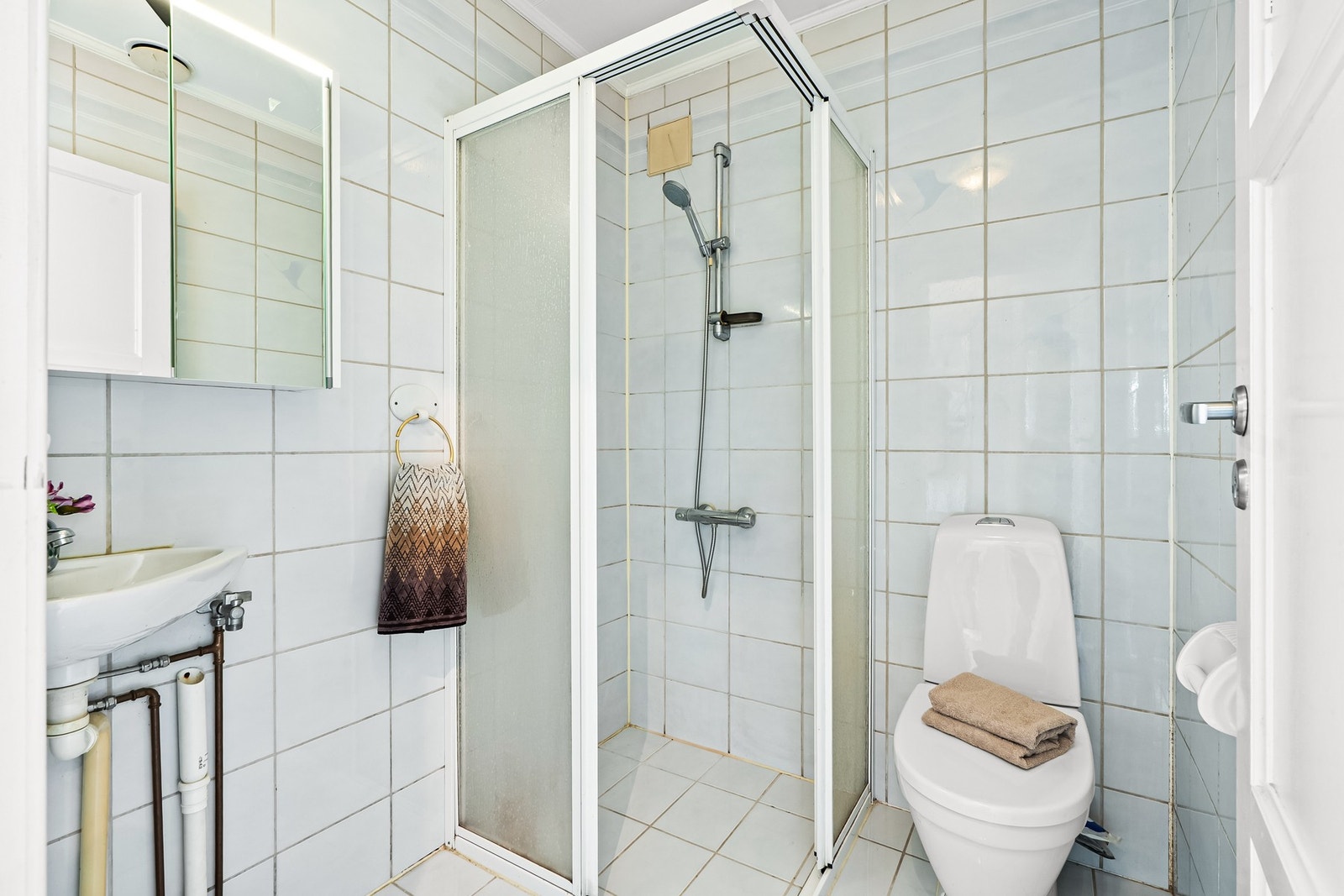 Badet har fliser på veggene og samme fliser på gulv med gulvvarme. Av innredning er det en hjørneservant og et speilskap og badet er videre utstyrt med et toalett, et dusjhjørne og opplegg for vaskemaskin. Galleribilde