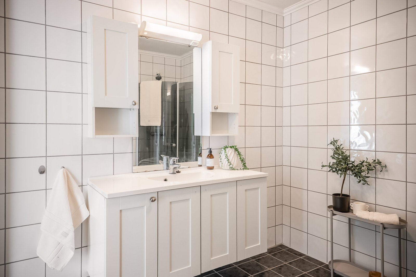 Flislagt bad/vaskerom med dusjhjørne med glassvegger, servantinnredning, gulvmontert toalett og opplegg for vaskemaskin. Galleribilde