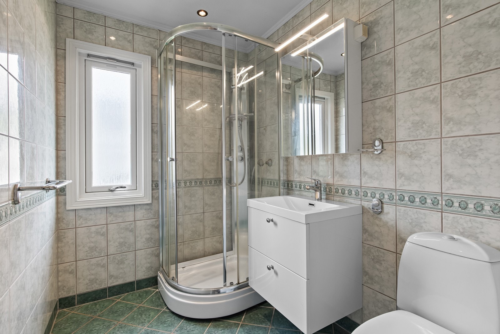 Badet er utstyrt med dusjkabinett, skuffseksjon med heldekkende servant, speilskap og toalett, og er flislagt på vegger og gulv. Galleribilde