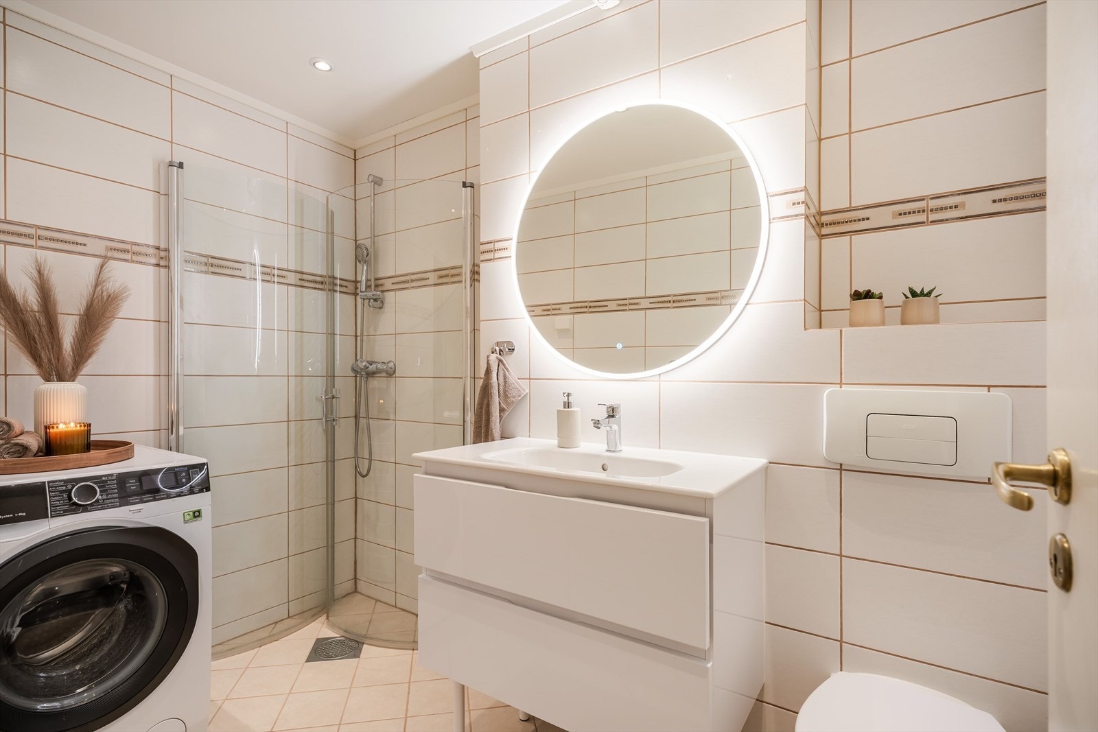 Badet består av gulv- og veggfliser, innfellbare dusjvegger av glass, vegghengt toalett, servantskap, lysspeil og opplegg for vaskemaskin. Galleribilde