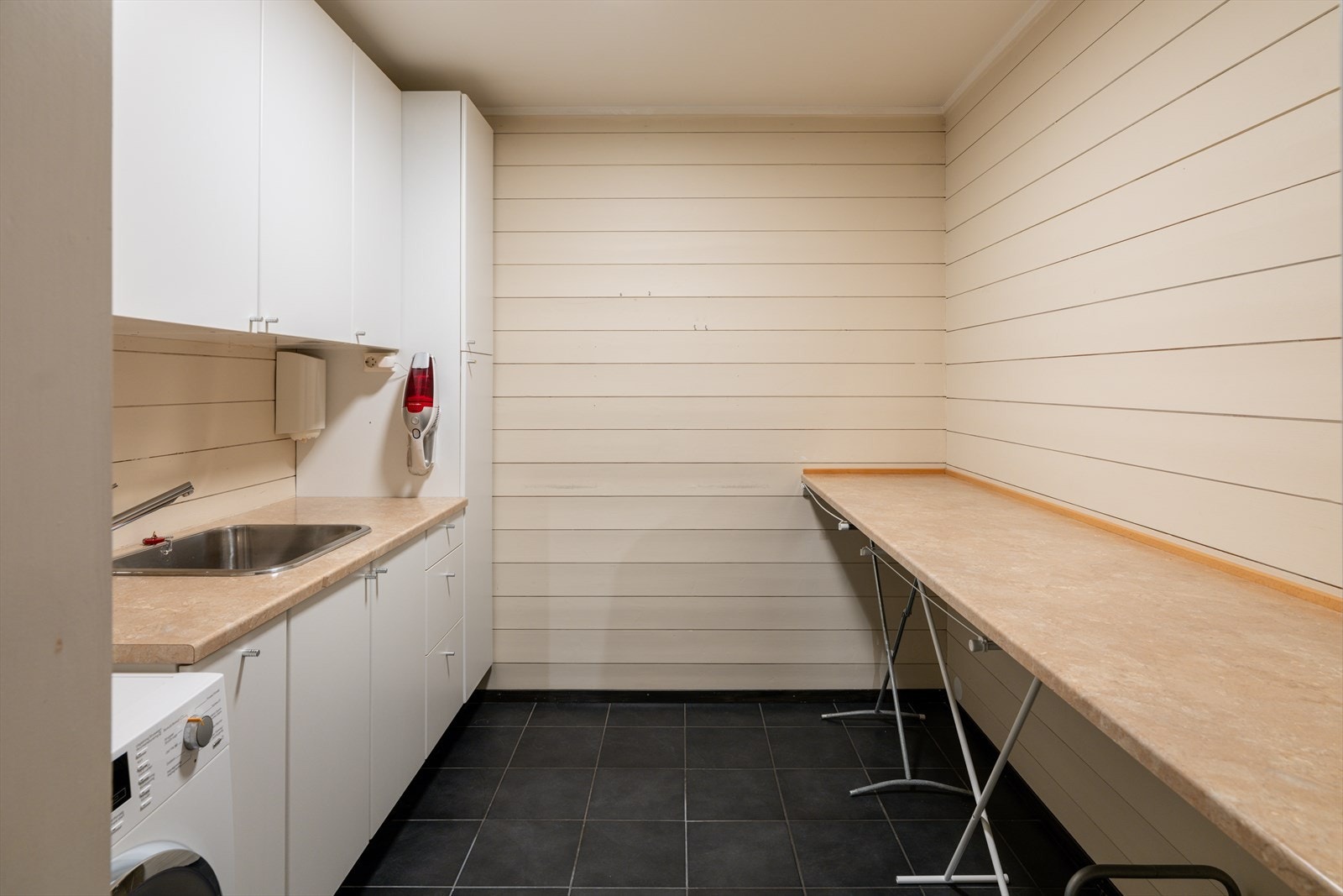 Praktisk innredet bod/vaskerom med skapinnredning, benkeplate med vask og opplegg for vaskemaskin og tørketrommel. Galleribilde