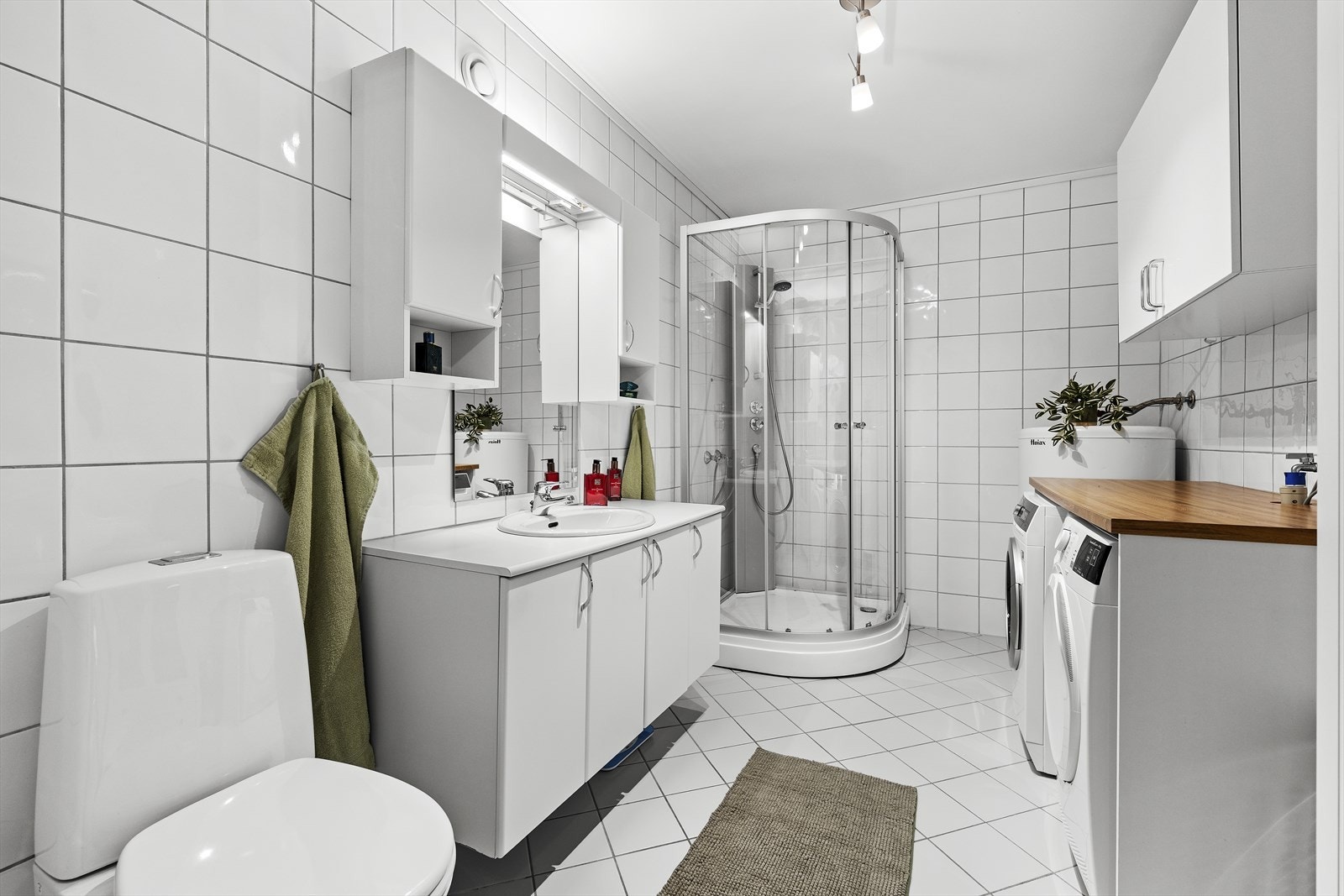 Lyst helfliset baderom med varmekabler i gulv. Badet er utstyrt med servant over skapinnredning, speil med overliggende belysning, sideskap, toalett, dusjkabinett og opplegg til vaskemaskin og tørketrommel Galleribilde