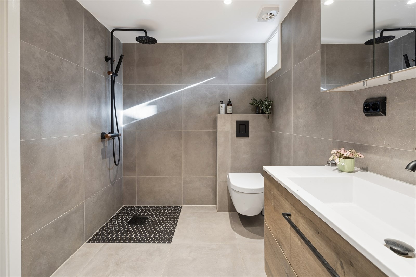 Badet i kjelleren har åpen dusjsone og regndusjsystem i sort. Vegghengt toalett. Moderne uttrykk med store grå fliser. Galleribilde